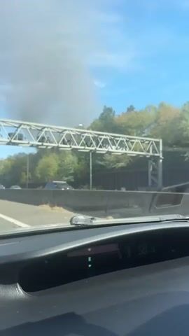 The video shows a view from inside a vehicle, likely a car, traveling on a highway. The dashboard display shows a speed of 58, then 57, then 56. In the distance, above a tree line and a concrete barrier, a plume of dark smoke is rising into the sky. Several other vehicles are visible on the highway, including cars and a white truck with "TRAWOGER" written on its side. The sky is blue with some wispy white clouds. The trees are green and some appear to be changing color, suggesting it might be autumn. A metal gantry with lights is visible over the highway. The person speaking is commenting on the smoke, stating it is not a car.