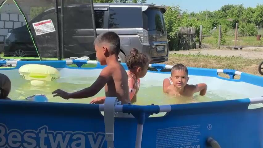 This is a video of three children playing in an inflatable swimming pool on a sunny day. The pool is blue and white, with the "Bestway" logo visible on the side. In the foreground, a young boy with dark hair and wearing white underwear is reaching out towards another child in the pool. In the middle, a young girl with blonde pigtails wears a pink life vest and plays with a yellow inflatable ring. To the right, a boy with dark hair smiles as he splashes in the water, also wearing a pink star-shaped floatation device on his arms. In the background, a gray minivan is parked, and beyond that, there is greenery and a wooden fence, suggesting a rural or suburban setting. The overall atmosphere is one of joyful summer recreation. The text visible on the pool reads "Bestway" and some smaller text which appears to be safety instructions or product information, though it is not clearly legible. The time of day appears to be midday, given the bright sunlight and shadows. The weather is clearly sunny and warm, ideal for swimming. The location context is Biysk, Russia.
