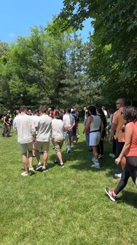 The video shows a group of people in a grassy outdoor area, likely a park or backyard, on a sunny day with blue skies and scattered clouds. A group of men and women are gathered around a long, narrow table, with some people standing on one side and others on the opposite side. The table appears to be set up for a game, possibly beer pong, as there are cups arranged on it. People are dressed casually in summer attire, including t-shirts, shorts, and hats. Some individuals are actively engaged in throwing objects towards the table, while others watch and socialize. The environment is lush and green, with trees providing shade in the background. The overall atmosphere suggests a social gathering or an outdoor party.