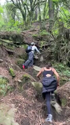 Two people hike on a steep, rocky, dirt trail in a forest. The person in front wears a dark jacket over a black tank top and black leggings. The person behind wears dark pants, a grey shirt, and a backpack. The forest has tall trees and mossy rocks and roots.