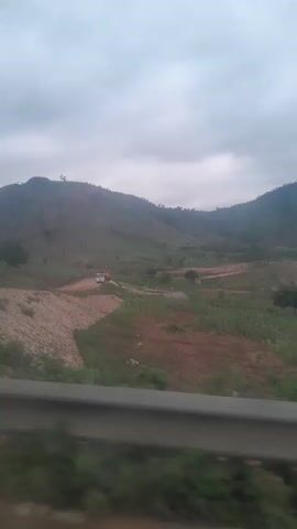 The video shows a landscape view from a moving vehicle. The landscape consists of rolling hills covered in vegetation, with a cloudy sky overhead. In the middle ground, there is a dirt road leading into a valley. There are a few small buildings or structures visible, as well as some construction or mining equipment, including what appears to be a truck. The vegetation includes green bushes and trees, as well as some cultivated fields, possibly corn. A signpost is visible on the left side of the road. The overall impression is of a rural or developing area with some ongoing construction.