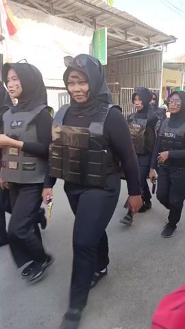 A group of young women, all wearing black hijabs and black clothing, are walking down a paved street. They are also wearing what appear to be tactical vests, some with writing on them. Several of the women are carrying what look like toy guns. The women appear to be part of a parade or event, possibly a costume contest. The background shows a street with buildings and some parked motorcycles. The overall scene suggests a playful or theatrical display rather than a serious event.