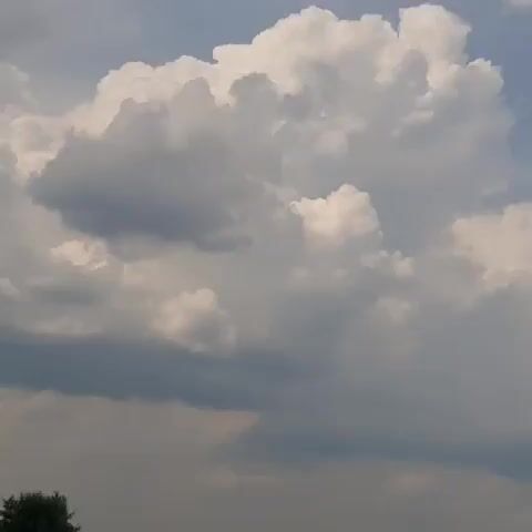The media file shows a dynamic sky filled with large, fluffy cumulus clouds. The lighting suggests it is daytime, likely late afternoon given the contrast between the bright white clouds and the darker, more ominous storm clouds gathering below. The lower portion of the frame reveals a hint of the ground, featuring a patch of green trees on the left and several white, tent-like structures in the distance. The presence of these structures and the distant trees indicates an outdoor setting, possibly a park, event space, or rural area. The overall mood conveyed by the sky is one of impending weather change, with the towering clouds suggesting potential for rain or a storm. There is no visible text in the media file. The location context of Dubrivka, Ukraine, places this scene in Eastern Europe.