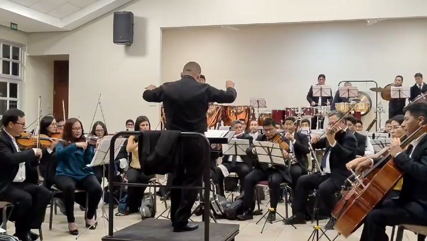 An orchestra is performing in a well-lit, simple room. The musicians are seated in rows, dressed in formal attire, playing instruments such as violins, cellos, and percussion. A conductor, also in formal wear, stands on a small podium facing the orchestra, directing them with broad arm movements. Sheet music is placed on stands in front of each musician.  A speaker is mounted high on the wall in the background. The room has neutral-colored walls and a white ceiling. A few bags are scattered on the floor near the musicians. There is a window on the left side of the frame.