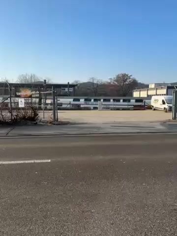 The video shows a street scene with a Hertz NeverLeave storage area in the background. The camera starts with a view of a gated area with several parked vehicles. The camera then pans to the right, showing the street and a person walking on the sidewalk. The sky is clear and blue. The overall environment appears to be an industrial or commercial area.