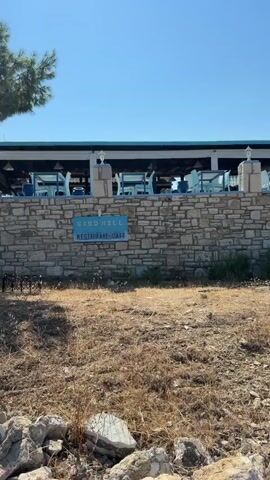 This is a video of an outdoor restaurant or cafe with blue tables and chairs. A stone wall is in the foreground, and a Greek flag is flying from a pole. The sky is clear and blue.