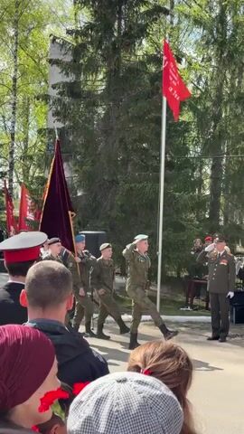 The video captures a parade or formal ceremony in an outdoor setting. A group of people is gathered, presumably as an audience, while four individuals in military uniforms, two with dark berets and two with light blue ones, are marching. One of the soldiers salutes a higher-ranking officer in a green military uniform standing on the sidelines. The soldiers are carrying a maroon-colored flag with a gold emblem and fringe, and another taller pole with the Russian flag is visible in the background. Behind the group are multiple flags on poles, including the Russian flag, a Soviet-era red flag, and what appears to be a regional or local flag with green and blue colors. The surroundings feature lush green trees, typical of a spring or summer day. The scene suggests an event commemorating military or national importance.