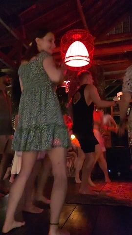 The video shows several people, mostly women, dancing and socializing in a dimly lit, club-like setting. The ambiance is created by red and purple lighting, with a prominent red, spherical lantern visible on the left side of the frame. People are moving to the music, some standing and actively dancing, while others are seated or moving more subtly. The setting appears to be a bar or lounge, possibly with wooden structures, consistent with a tropical or rustic venue. The time of day is clearly night, indicated by the artificial lighting. There is no visible text. The atmosphere is lively and festive, suggesting an enjoyable gathering in Lokaserena, Indonesia.