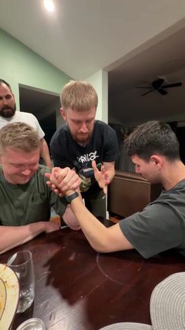 Here's a detailed description of the video:

The video shows four men in a domestic setting, around a dark brown wooden table. Two of the men are engaged in an arm-wrestling match. The man on the left has short, light brown hair and wears a dark green short-sleeved t-shirt. He is visibly straining with a strained expression on his face. The man on the right has dark hair, a mustache, and a small earring in his left ear; he is wearing a gray t-shirt. Both men have their right elbows on the table and their hands tightly clasped, wrestling.

A third man stands between them, observing and moderating the arm wrestling match. He has short red hair, a beard and wears a black t-shirt with a graphic. He is smiling slightly, with his hands up in the air.

The fourth man is standing behind the arm wrestlers, closer to the background. He has dark hair, a beard, and wears a white t-shirt. He has his right hand up near his mouth and is smiling.

The table is cluttered with items, including empty dishes, glasses, and a silver can. The room is brightly lit with a ceiling light. The walls are painted in shades of green and white. There's a ceiling fan in the background. In the background, a brown leather couch is visible.

The atmosphere is friendly and energetic. The men are focused and engaged in the arm-wrestling match.
