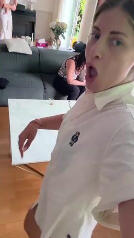 A woman with blonde hair, wearing a white button-down shirt with a bear logo, is filming herself in a selfie video. She makes various facial expressions, including sticking her tongue out and pouting. In the background, another woman with dark hair is sitting on a couch, looking at her phone. A coffee table with a white marble top is in the foreground, and a dark couch is visible behind it. The environment appears to be a living room with wooden floors and large windows. The video has a slightly shaky camera motion.