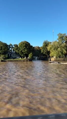 The video shows a river with brown, murky water flowing past the camera. On the riverbanks, there are buildings and trees. A small motorboat speeds along the river, creating a wake. The sky is blue and cloudless. The environment appears to be a riverfront area with houses or buildings along the shore.