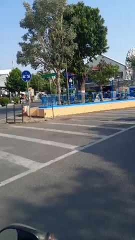 The video shows a street scene with a blue minivan and a black SUV. The minivan is in the foreground, with its rear facing the camera. The SUV is to the right of the minivan, also facing away from the camera. The street is paved and has white lines marking lanes. There are trees and buildings in the background. The sky is clear and blue. There is a Ferris wheel in the background. There are also some orange traffic cones and a white sign with graffiti on it. The overall scene appears to be a sunny day in a town or city.