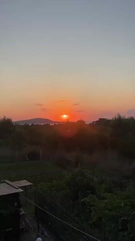 The video shows a sunset over a rural landscape. The sun is setting behind a hill in the distance, casting a warm orange glow over the scene. There are trees and foliage in the foreground, and a body of water can be seen between the hill and the trees. The sky is a gradient of orange, pink, and blue, with a few scattered clouds. The video appears to be shot from a slightly elevated perspective, possibly a balcony or a hill. There are no people or animals visible in the video.