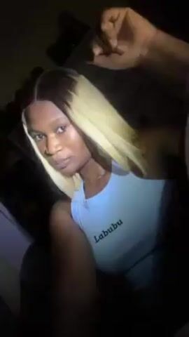 A young woman with dark skin and a blonde wig is facing the camera. She is wearing a light blue tank top with "Labubu" printed on it. She poses by placing her hand under her chin. The lighting is poor, and the background shows a dark room with some shelving.