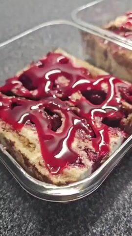The video shows a close-up, side-angle view of three square clear plastic containers, each filled with slices of a dessert. The dessert appears to be a fruit crumble or pie with a golden-brown base and a topping of sliced red fruit, possibly cherries or berries, coated in a dark red glaze or sauce. The containers are placed on a gray, speckled countertop. The first container in the foreground is not covered. The second and third containers, further back and to the right, are covered with clear plastic wrap. The video pans slightly to the right, revealing more of the dessert in the containers and the texture of the countertop.