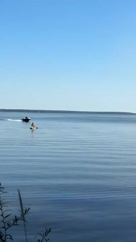 A video captures a wide shot of a calm lake on a clear day. The horizon is flat and distant, with a faint line of trees. The sky is a solid, bright blue with no clouds. In the foreground, the water is rippling gently, creating horizontal lines of light and shadow. On the water, a small green and white motorboat is moving from left to right, leaving a slight wake. To the left of the motorboat and slightly closer to the viewer is a person on a green paddleboard. The person is wearing a red top and appears to be paddling. The video is static, with no camera movement.