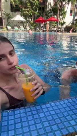 A young woman in a dark swimsuit is in a swimming pool, holding and drinking from a clear plastic cup with a green straw. The cup appears to contain orange juice or a similar bright-colored drink.  She is looking directly at the camera with a slight smile. A person swims past her in the background, wearing blue swim trunks and holding a colorful ball. The pool has blue tiles and other people are visible swimming or relaxing on lounge chairs in the background. The environment appears to be a resort or hotel swimming pool due to the presence of umbrellas, trees, and a multi-story building.