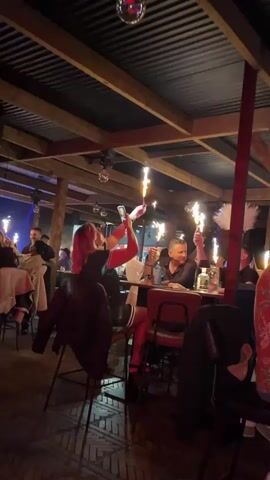 The video shows a group of people celebrating at a bar or club with sparklers. A man and woman are seated at a table, each holding up a sparkler, while another man sits nearby. Other people are seated at nearby tables. A waiter passes by. The space has a rustic feel with wooden beams and a corrugated metal ceiling. Disco balls and blue lights add to the festive atmosphere.