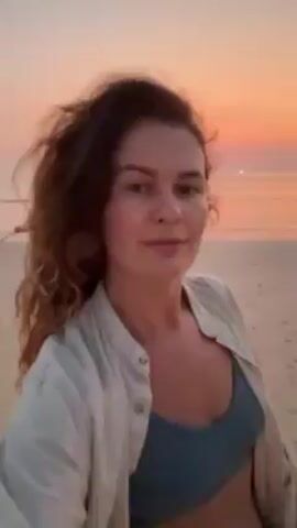 A woman stands on a beach, facing the camera with a slight smile. She has brown, curly hair and is wearing a light-colored, open button-down shirt over a blue sports bra. The background shows a beach with gentle waves coming ashore, and a sunset sky displaying shades of orange and pink. The scene appears peaceful and serene.