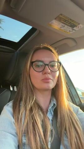 A young woman with long blonde hair and glasses is sitting in the driver's seat of a car. The sun is shining through the sunroof, casting a warm glow on her face and hair. She is wearing a light blue denim shirt. The interior of the car is visible, including the dashboard and steering wheel. The woman appears to be speaking or singing, as her mouth is slightly open. The overall scene suggests a candid moment captured while driving.
