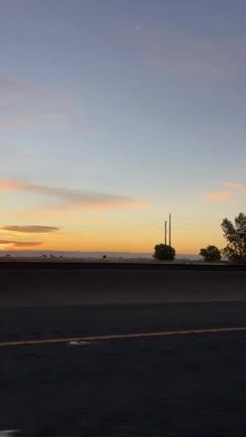 The video shows a view from a vehicle traveling along a highway at dusk. The sky is a gradient of pale blue fading into orange and pink hues, indicative of a sunset or sunrise. A sliver of the moon is visible in the upper right corner of the sky. Along the horizon, silhouetted trees and power lines are present. A concrete barrier runs along the side of the highway in the foreground, with a double yellow line marking the edge of the road. The overall scene suggests a peaceful, transitional moment during travel.