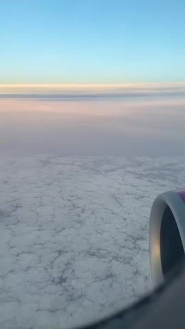 The video shows a view from an airplane window. Below the aircraft is a vast expanse of white clouds, resembling a textured blanket. Above the clouds, there is a band of hazy pink and orange colors merging into the clear blue sky at the top of the frame. Part of the plane's engine nacelle is visible in the bottom right corner.