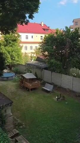 A drone shot shows a backyard with a small blue pool, a wooden playhouse with a slide, and a swing set. A concrete fence separates the yard from neighboring properties. Behind the fence, there are buildings with red tile roofs and a white facade. Trees with green foliage are visible throughout the scene, and the sky is partly cloudy. The camera moves slightly, revealing more of the surrounding buildings and a street with a parked car. The video appears to be shot during the day.