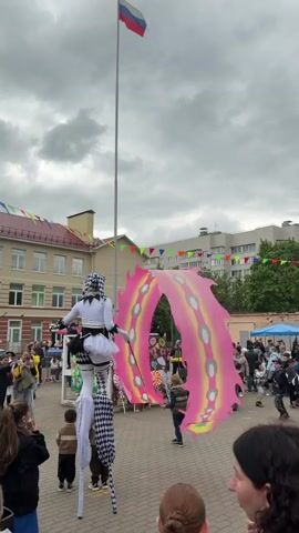 The video shows a crowd of people in an outdoor public space, possibly a town square or a fairground.  In the foreground, a person dressed in a white and black checkerboard-patterned costume and wearing stilts stands out. This individual also holds a decorative wand. Next to them, two people walk with a large, brightly colored pink and yellow butterfly-shaped decoration. In the background, there is a school building adorned with festive pennants. A Russian flag flies prominently from a tall flagpole. The sky is overcast with grey clouds. Other buildings and trees are visible in the surrounding area. People are walking around, some taking pictures, indicating the presence of a community event or festival.