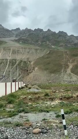 This is a vertical video of a mountainous landscape with a river flowing through a valley. The sky is overcast with grey clouds. In the foreground, there is a rocky, dirt path with some green vegetation. In the middle ground, a river flows from right to left, with grassy slopes on either side. The slopes are steep and have areas of exposed rock and scree. In the background, jagged, rocky mountain peaks rise against the cloudy sky. One of the peaks has a dusting of snow. There is a signpost to the left of the frame which appears to be an advertisement. The video is filmed from a moving vehicle.