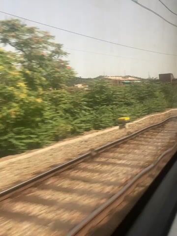 The video shows a view from a moving train traveling along a railroad track. The track itself is visible in the foreground, with ballast and sleepers. To the left of the track, there is a landscape with green bushes and trees, as well as some buildings in the distance. The sky is clear and bright, suggesting a sunny day. There are some overhead power lines visible, indicating that this is an electrified railway. The buildings in the background appear to be residential structures, and some have reddish-brown roofs. The terrain seems somewhat hilly. The motion blur indicates the train is moving at a moderate speed. There are no people visible in the frame, and no specific objects or actions stand out beyond the general scene of a train journey through a semi-urban or rural environment.