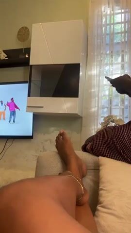 Here's a description of the video:

The video shows a person lounging on a couch while watching a music video on a TV. The TV is mounted on a wall above a fur-covered entertainment center. There's a white cabinet mounted on the wall to the right of the TV.  The person's legs are visible, and they're wearing an anklet. The living room has light green walls, a wooden door, and white curtains. A glimpse of a kitchen area is also visible.