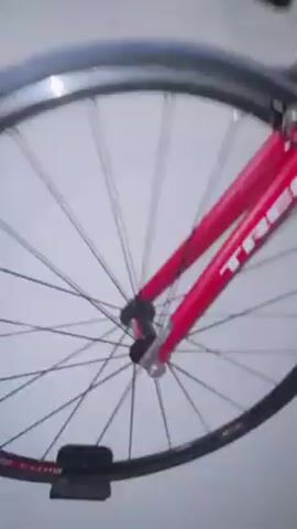 The video shows a red Trek bicycle with white lettering. The camera moves around the bike, showing the front wheel, frame, and handlebars. The background is a plain, light-colored wall. The video appears to be taken indoors. There is no one visible interacting with the bike.