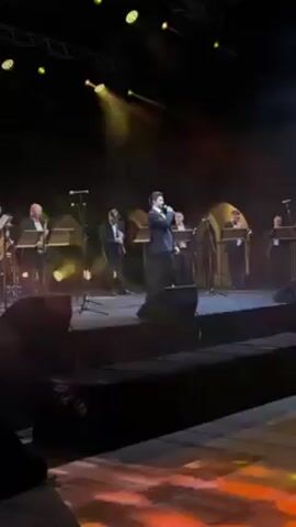 On a stage illuminated by yellow spotlights, a man in a black suit sings into a microphone. Behind him stand six musicians, also in suits, playing various instruments. The musicians are positioned behind ornate music stands. The stage appears to be part of a larger venue, evidenced by the overhead rigging and lighting. The overall atmosphere suggests a live performance.