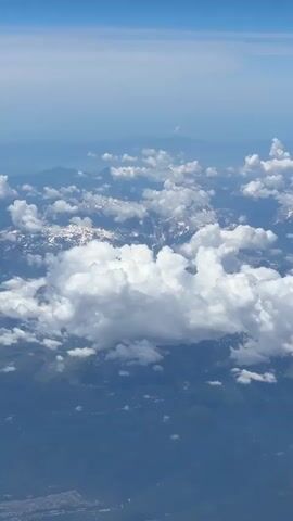 The video shows an aerial view from an airplane, looking down at a mountainous landscape partially covered by clouds. The sky above is blue with some wispy clouds. Below, mountain peaks are visible, some with snow caps. Large, fluffy white clouds are scattered across the scene, casting shadows on the mountains. The lower part of the frame shows green terrain with hints of human settlements, possibly towns or villages. The view suggests the airplane is flying at a considerable altitude.