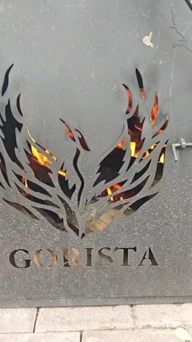 The video shows a close-up, static shot of a metal grill with a design cut into it. The design appears to be a bird, possibly a phoenix, with flames behind it. The word "GORISTA" is cut into the metal below the design. There is a latch visible on the right side of the grill. The flames are visible through the cutouts in the metal. The grill is outdoors, as indicated by the brickwork at the bottom of the frame. The lighting suggests it is daytime, possibly evening due to the presence of flames.
