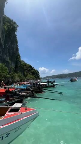 The video shows a body of turquoise water with several boats docked near a cliff face. On the left side of the frame, a white and red speedboat is in the foreground. Two individuals are visible on this boat. Further back, a line of traditional Thai longtail boats are moored. People are seen on the shore and on the boats, some appear to be tourists. The cliff face is covered in lush green vegetation. The sky is blue with scattered clouds. The water is clear and calm.