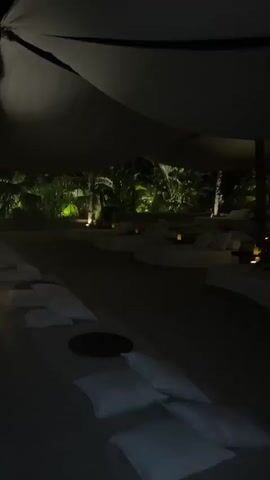 The video shows an outdoor seating area at night. The area is covered by a large, dark canopy. In the foreground, there are several white cushions scattered on a dark surface. In the middle ground, there are several round, white seating pods arranged in a semi-circle, each with a small candle on it. Behind the seating pods, there is a dense line of tropical foliage, illuminated by spotlights that cast a green glow. The lighting creates a moody and atmospheric scene.