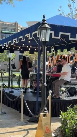 A female singer in a black sequined dress performs on a stage in front of a fountain and a large golden statue. She is backed by a band consisting of a keyboardist, a drummer, and another musician playing a guitar or bass. The stage is covered by a blue awning with decorative trim and lights. A street lamp stands in the foreground, and a "Caution Wet Floor" sign is visible at the bottom right. The setting appears to be an outdoor plaza with shops in the background.