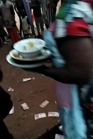 The video shows a low-angle, somewhat shaky, indoor scene with music playing. Initially, the view is obscured by what appears to be a brightly patterned piece of clothing. The camera shifts to reveal a dark floor with scattered white rectangles, possibly paper or currency. The view then moves towards a person wearing a light blue suit and black shoes, playing a saxophone. The person is partially obscured by musical equipment and other items. The scattered white rectangles are visible around the musician. The video ends with the person continuing to play the saxophone.