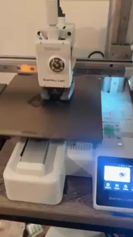 The video shows a Bambu Lab 3D printer in operation. The white printer head moves along a track, applying a layer of material onto a dark grey print bed, gradually forming a shape. The printer's control panel shows temperature readings. The device sits on a wooden surface in what appears to be an indoor setting.