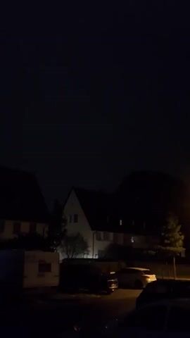 The video shows a dark outdoor scene at night. In the foreground, a portion of a dark-colored car is visible. In the mid-ground, several parked cars are dimly lit. Beyond the cars, two residential buildings with illuminated windows are visible. A streetlamp with a bright yellow light is also present in the mid-ground, casting light on the surroundings. Trees are also visible, silhouetted against the night sky. The sky itself is dark, indicating nighttime. The camera appears to be handheld and is slightly shaky, causing some motion blur, especially when the streetlamp is in view. There are no people clearly visible in the frame.