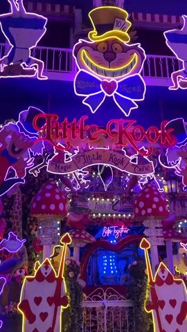 The video displays the exterior of a cafe decorated with an Alice in Wonderland theme. Neon lights in various colors outline figures and lettering. In the foreground, two large card figures, the Queen of Hearts, stand symmetrically on either side of an entrance. Above the entrance, the cafe's name, "Little Kook Cafe," is displayed in neon. To the left, a depiction of the Cheshire Cat with a wide grin is visible. Red and white mushroom figures are also integrated into the decoration. The overall scene is vibrant and fantastical, illuminated by the bright neon signs against a dark background.