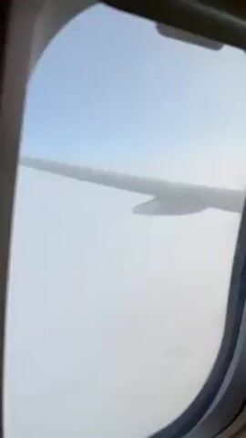 The video shows a view from inside an airplane, looking out the window. The airplane wing is visible outside the window. The sky is light blue, but there is a thick layer of white clouds below the plane.