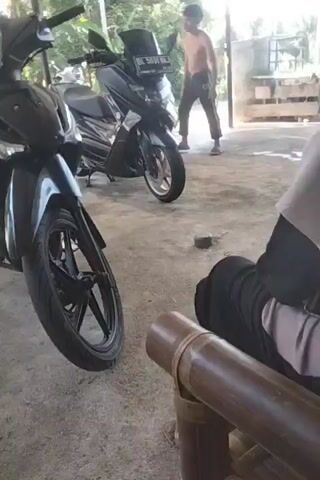 The video begins with a view of three motorcycles parked in a covered outdoor area. A shirtless man stands near one of the motorcycles. The camera shifts perspective, focusing on the legs and feet of a person walking near a white scooter and another motorcycle. The camera is stationary and positioned low, likely held by someone sitting. The scene appears to be an outdoor garage or carport with sunlight shining in. The ground is concrete and somewhat dusty. Other objects visible include a broom, some concrete blocks, and various trees in the background.
