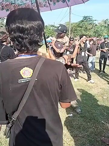 The video shows a group of people at an outdoor event. The group consists of men, one of whom is carrying a child on his shoulders. They are standing on a grassy area under a large umbrella, possibly for shade. Most of the men are wearing casual clothes, including black t-shirts, jeans, and baseball caps. Some of them have lanyards around their necks. The child on the man's shoulders is also wearing a baseball cap. Other people are visible in the background, along with some tents and equipment, suggesting this may be a fair or community event. The people in the foreground are talking and laughing, indicating a social atmosphere. One of the men is holding the umbrella to provide shade for the group.