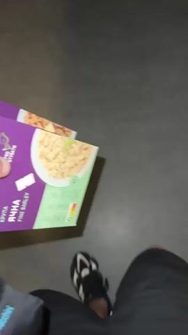 The video shows a person walking indoors carrying two boxes of fine barley. The person's legs are visible, wearing black shorts and black and white sneakers with a green accent. The boxes of barley are purple, green, and white, with a picture of cooked barley on the front. The floor appears to be a solid gray color. The person is moving forward.