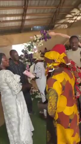 In what appears to be an indoor event space with a corrugated metal roof, a group of people are gathered around a floral archway. The archway is adorned with white and purple flowers. Among the people, several are wearing traditional African print clothing, including yellow, red, and brown patterns. There's also a person in a white lace dress. They appear to be dancing and interacting, possibly at a wedding reception or similar celebration.