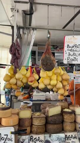 A stall is set up with various food items. In the foreground, several rounds of cheese are stacked and displayed, some with grated cheese on top. Behind the cheese, bunches of yellow, pear-shaped cheeses hang from the ceiling, suspended in netting. To the left, strings of dark red sausages hang. In the center, a large, cured ham is suspended. To the right, a whiteboard lists "NOSTRI FRESCHI," which translates to "OUR FRESH," followed by a list of cheeses including "BURRATA," "MOZZARELLA DI BUFALA," and "RICOTTA." Behind the stall, a person in a red shirt is seen working, and other people and shelving units are visible in the background. The stall appears to be outdoors or in a market setting.