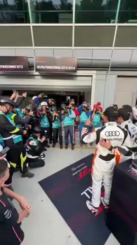 The video captures a vibrant post-race scene at a pit lane in Monza, Italy. Two race car drivers, one wearing a white, black, and orange racing suit with "Audi Sport" and "San Lorenzo" logos, are standing proudly on a black mat, posing with two large trophies placed on black pedestals. They are surrounded by a throng of photographers and videographers, many kneeling to get the best shot, all pointing cameras towards the drivers.

In the background, a modern building with large glass windows and garage doors is visible, with signs overhead reading "SCRUTINEERING GT3" and "SCRUTINEERING GT2/GT4." A black Audi R8 LMS GT3 race car, featuring red accents and sponsor decals such as "Pirelli," "Castrol," "OKX," and "APEX," is parked nearby. Spectators are gathered in the background, observing the celebration. The setting is bright and sunny, indicative of daytime. A partial view of a "Pirelli" banner can be seen in the distance, further confirming the race track environment.