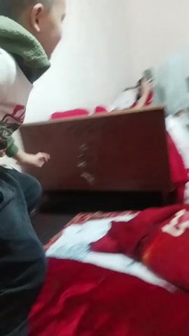 A young boy dressed in dark blue sweatpants, a white long-sleeved shirt with a green hood, and dark socks attempts to climb on top of a wooden headboard located at the end of a bed covered with a red and white patterned blanket. He pulls himself up, smiles, and stands on the headboard. Behind him, someone appears to be lying in the bed, looking at a phone. The room has white walls.